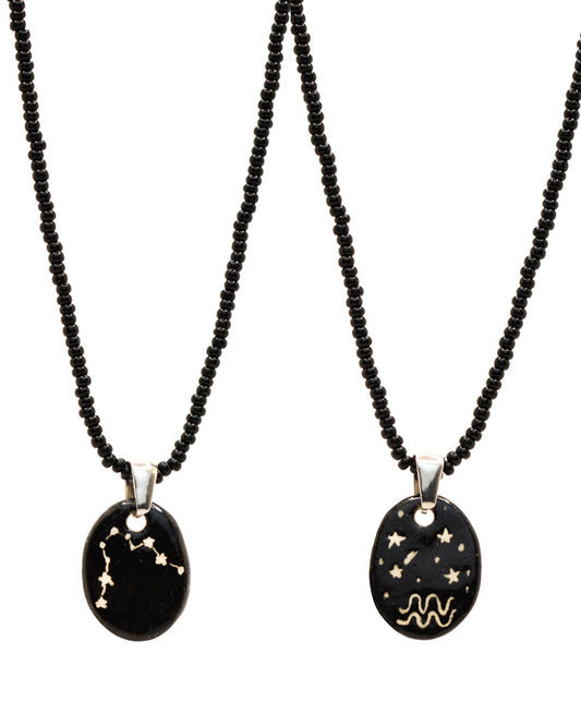 Colar elegante com pendente de cerâmica dupla-face representando a constelação Aquário, adornado com missangas de vidro pretas para um toque sofisticado e misterioso.