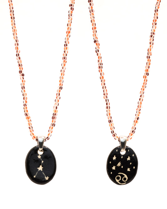Colar elegante com pendente dupla-face esculpido em cerâmica, retratando a constelação de Caranguejo, complementado por missangas listradas em tons castanhos para um toque de estilo e charme distintos.