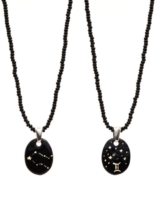 Colar elegante com pendente de cerâmica dupla-face representando a constelação Gémeos, adornado com missangas de vidro pretas para um toque sofisticado e misterioso.