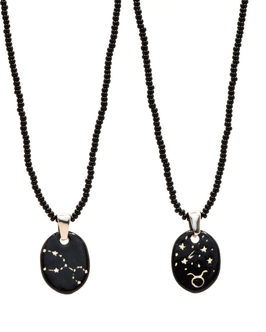 Colar elegante com pendente de cerâmica dupla-face representando a constelação Touro, adornado com missangas de vidro pretas para um toque sofisticado e misterioso.