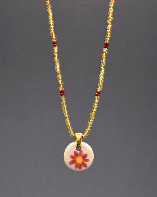Pendente encantador de margarida vermelha em cerâmica, meticulosamente pintado à mão, suspenso com graciosidade por um colar de missangas de vidro douradas semitransparentes, adornadas com um elegante padrão de bordô. Uma fusão única de arte e moda, este acessório acrescenta um toque floral vibrante e um brilho sutil ao seu estilo diário.