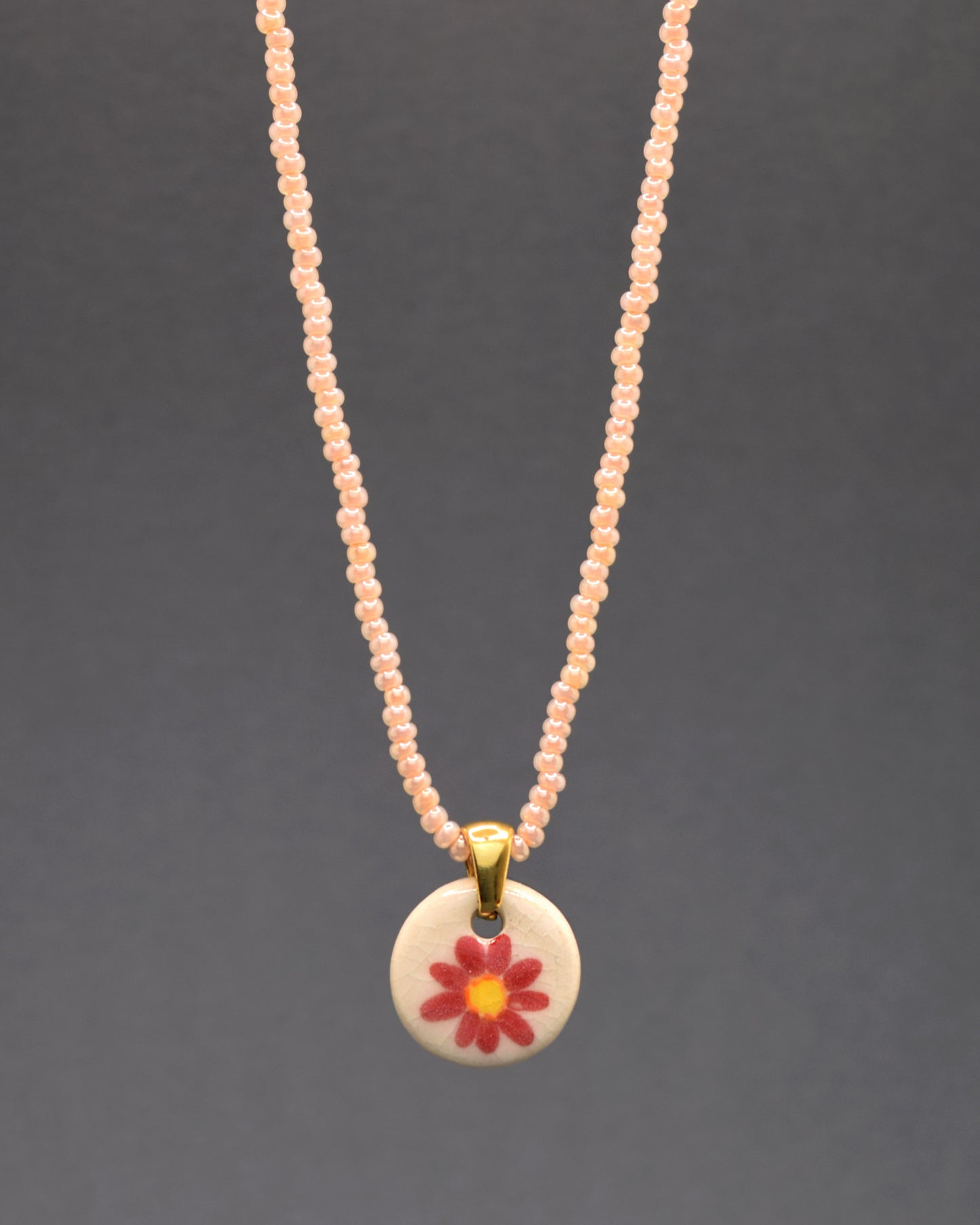 Pendente encantador de margarida vermelha em cerâmica, meticulosamente pintado à mão, suspenso com graciosidade por um colar de missangas de vidro salmão. Uma combinação delicada de arte e estilo, este acessório acrescenta um toque floral vibrante e uma sutileza colorida ao seu visual diário.