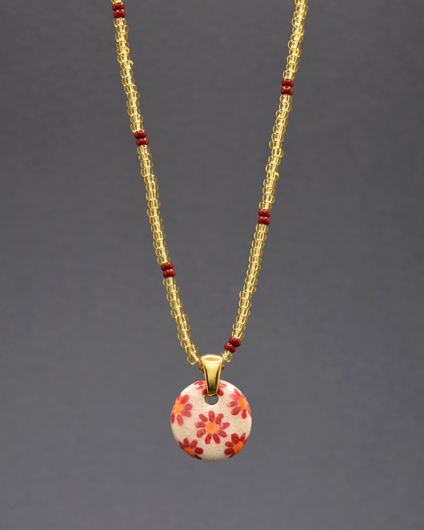 Explore a elegância única deste colar, onde um pendente de cerâmica com fundo branco e margaridas vermelhas é delicadamente suspenso em missangas douradas semitransparentes com padrão bordô. Uma fusão encantadora de artesanato refinado e design moderno, este colar emana sofisticação, adicionando um toque de brilho dourado e tons ricos ao seu estilo.