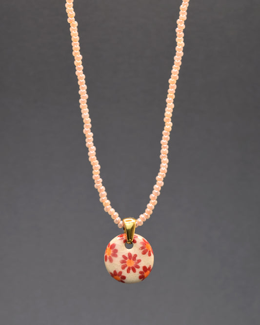 Este cativante colar exibe um pendente de cerâmica com fundo branco, realçado por vivas margaridas vermelhas pintadas à mão. Suspenso por um colar de missangas salmão, a peça transmite uma atmosfera encantadora, unindo o artesanato delicado ao vibrante toque de cor. Uma escolha elegante para adicionar um brilho distinto ao seu visual diário.