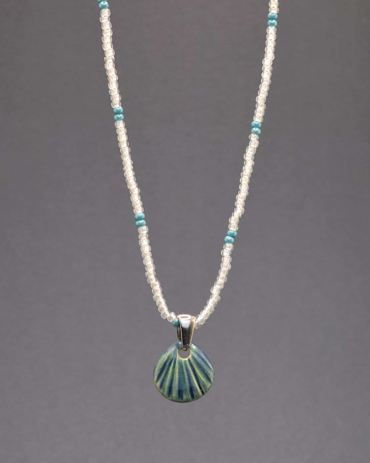 Eleve o seu visual com este colar de missangas de vidro brancas semitransparentes com padrão turquesa, adornado com um pendente de cerâmica artesanal em forma de concha verde musgo. Cada detalhe captura a elegância e serenidade da natureza, tornando esta peça uma expressão única de arte e beleza.