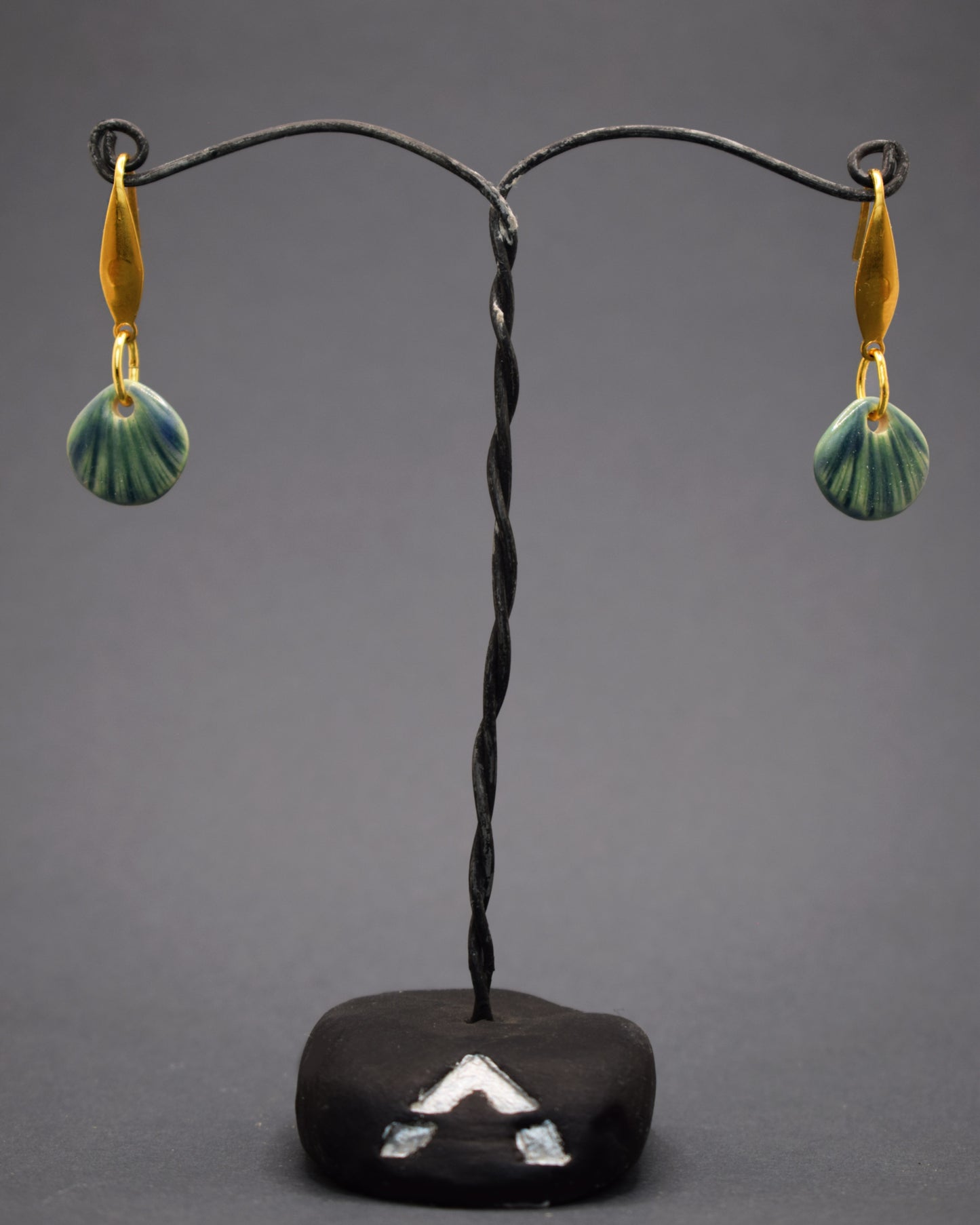 Brincos em aço inoxidável dourado com pendentes de cerâmica em forma de concha, na cor verde musgo. Combinando a durabilidade do aço com a delicadeza da cerâmica artesanal, estes brincos trazem um toque natural e único ao seu visual.