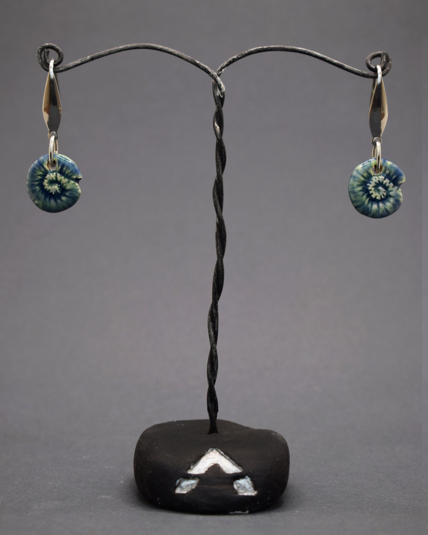 Adicione um toque natural ao seu estilo com brincos de aço inoxidável prateado, cada um com um pendente de cerâmica feito e pintado à mão, exibindo um caracol verde musgo. Uma combinação única de arte e natureza para um visual encantador.