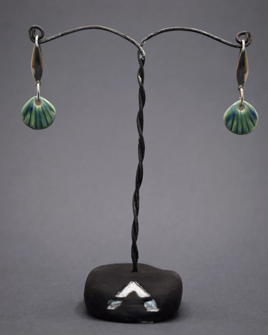 Brincos em aço inoxidável prateado com pendentes de cerâmica em forma de concha, na cor verde musgo. Combinando a durabilidade do aço com a delicadeza da cerâmica artesanal, estes brincos trazem um toque natural e único ao seu visual.