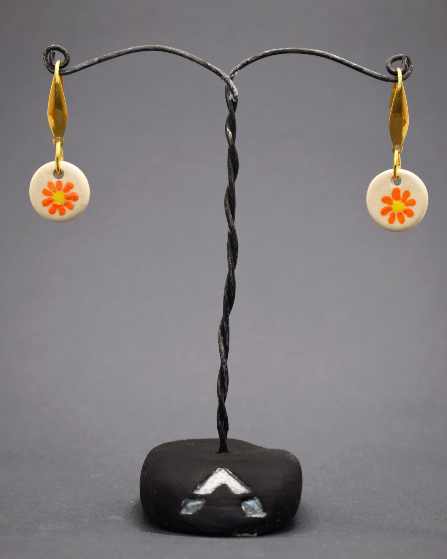 Realce seu estilo com brincos de aço inoxidável dourado, cada um com um pendente de cerâmica pintado à mão exibindo uma margarida laranja vibrante. Um toque de cor e frescor que celebra a beleza natural e a arte artesanal.