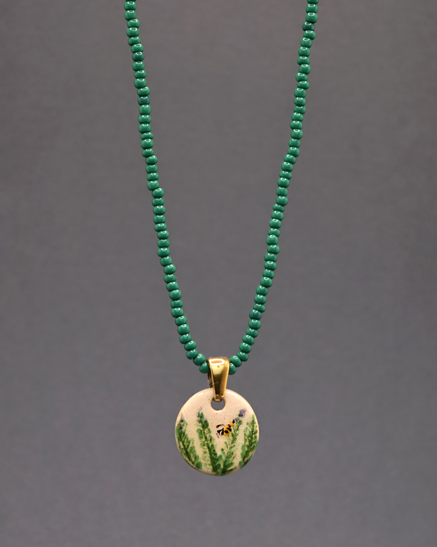 Colar encantador com missangas de vidro verdes e um pendente de cerâmica pintado à mão. O design exibe graciosos ramos de alecrim e uma abelha, adicionando uma dose de frescor e natureza à peça. Uma joia que une simplicidade e elegância num toque vibrante.