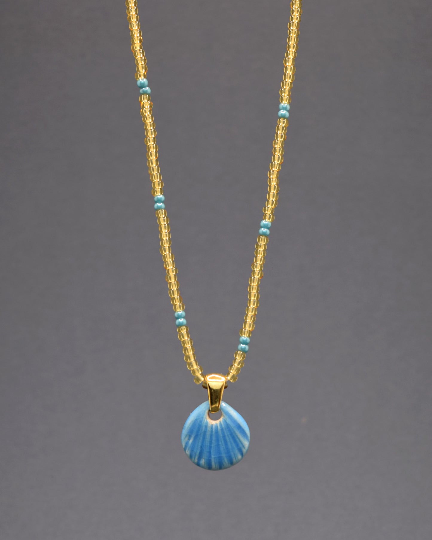 Descubra este colar de missangas de vidro douradas semitransparentes com padrão turquesa, adornado com um pendente de concha em cerâmica azul clara. Com um design artesanal e delicado, ele adiciona um toque de elegância e sofisticação ao seu visual, perfeito para qualquer ocasião.