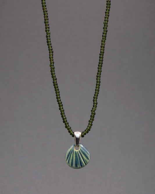 Encante-se com este colar de missangas de vidro verde escuro semitransparentes, adornado com um pendente de cerâmica artesanal em forma de concha verde musgo. Uma peça única que captura a serenidade e a elegância da natureza em cada detalhe.