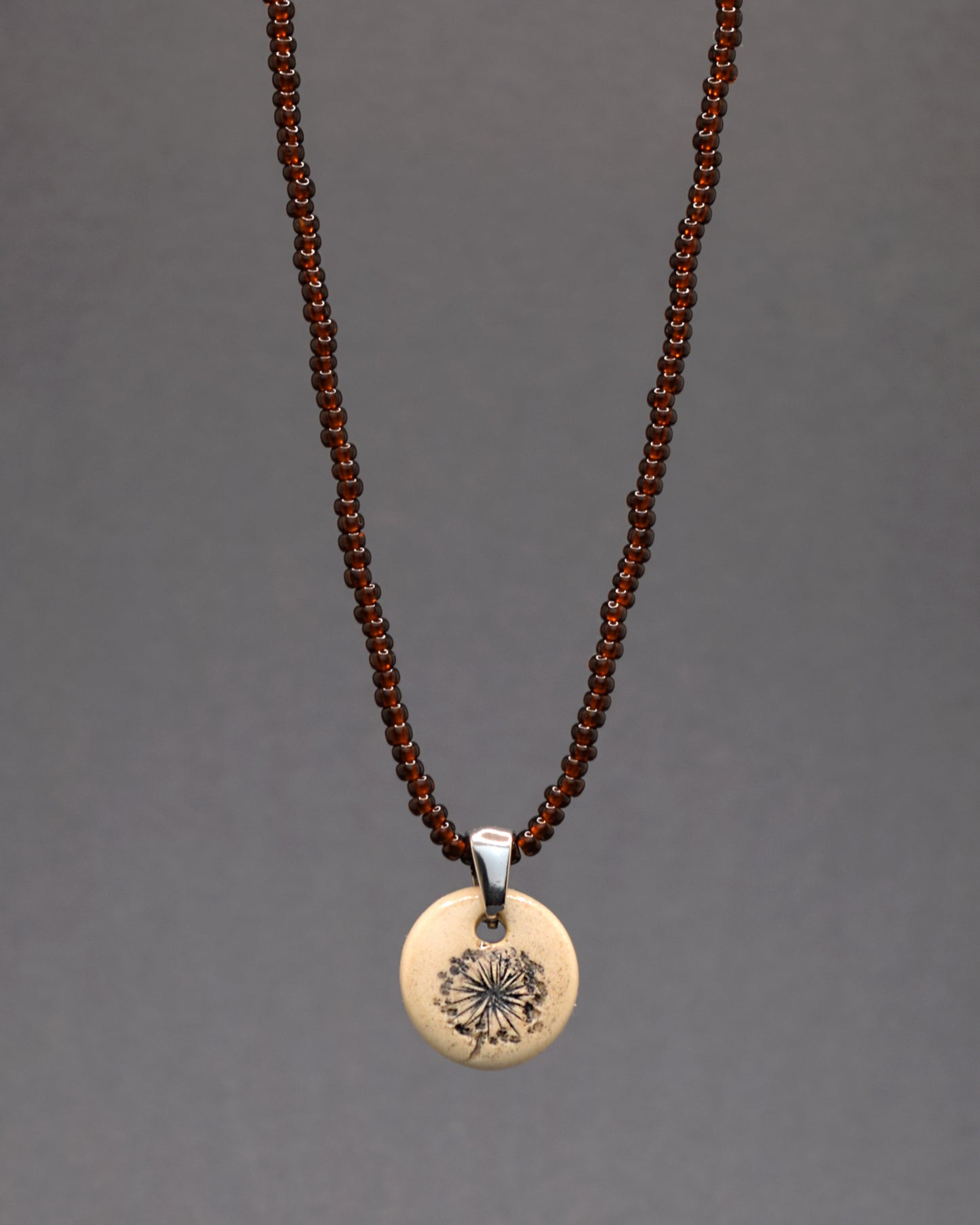 Este colar único, composto por missangas de vidro castanhas semitransparentes, é realçado por um pendente de cerâmica feito à mão, delicadamente adornado com o desenho de plumas de dente de leão. A combinação das cores terrosas com o simbolismo das plumas oferece uma peça cheia de charme e significado, perfeita para quem valoriza a beleza natural e a arte delicada no seu quotidiano.