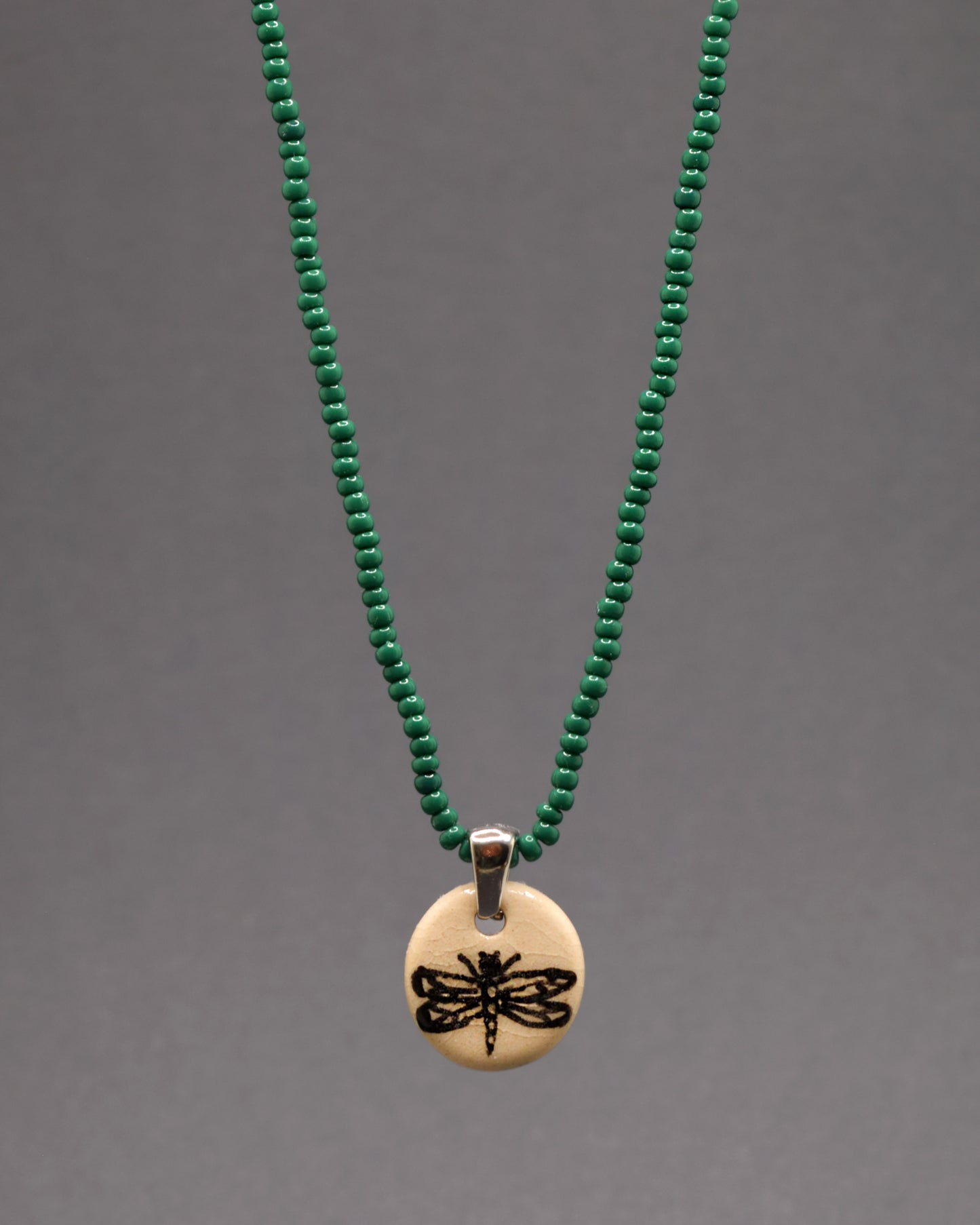 Colar de missangas de vidro verde com pendente de cerâmica, adornado com uma libélula desenhada à mão. A combinação vibrante das missangas com o toque artístico do pendente faz deste acessório uma peça original e elegante, perfeita para realçar qualquer look com charme.