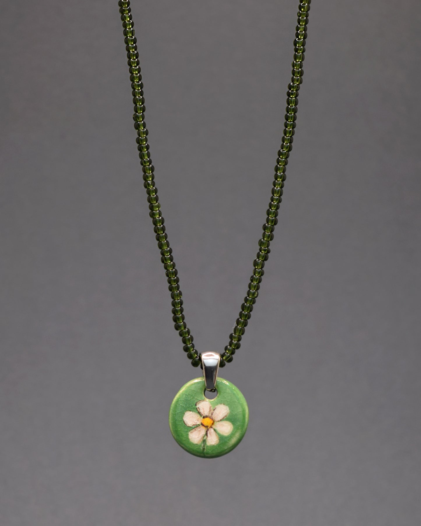 Explore a elegância discreta deste colar, composto por missangas verde escuro semitransparentes. As missangas formam uma peça sutil e refinada, enquanto o pendente de cerâmica, sobre um fundo verde, exibe uma margarida pintada à mão, proporcionando um toque artesanal encantador. Uma escolha distinta que combina a modernidade das missangas com a autenticidade da cerâmica, elevando o seu estilo com simplicidade e sofisticação.