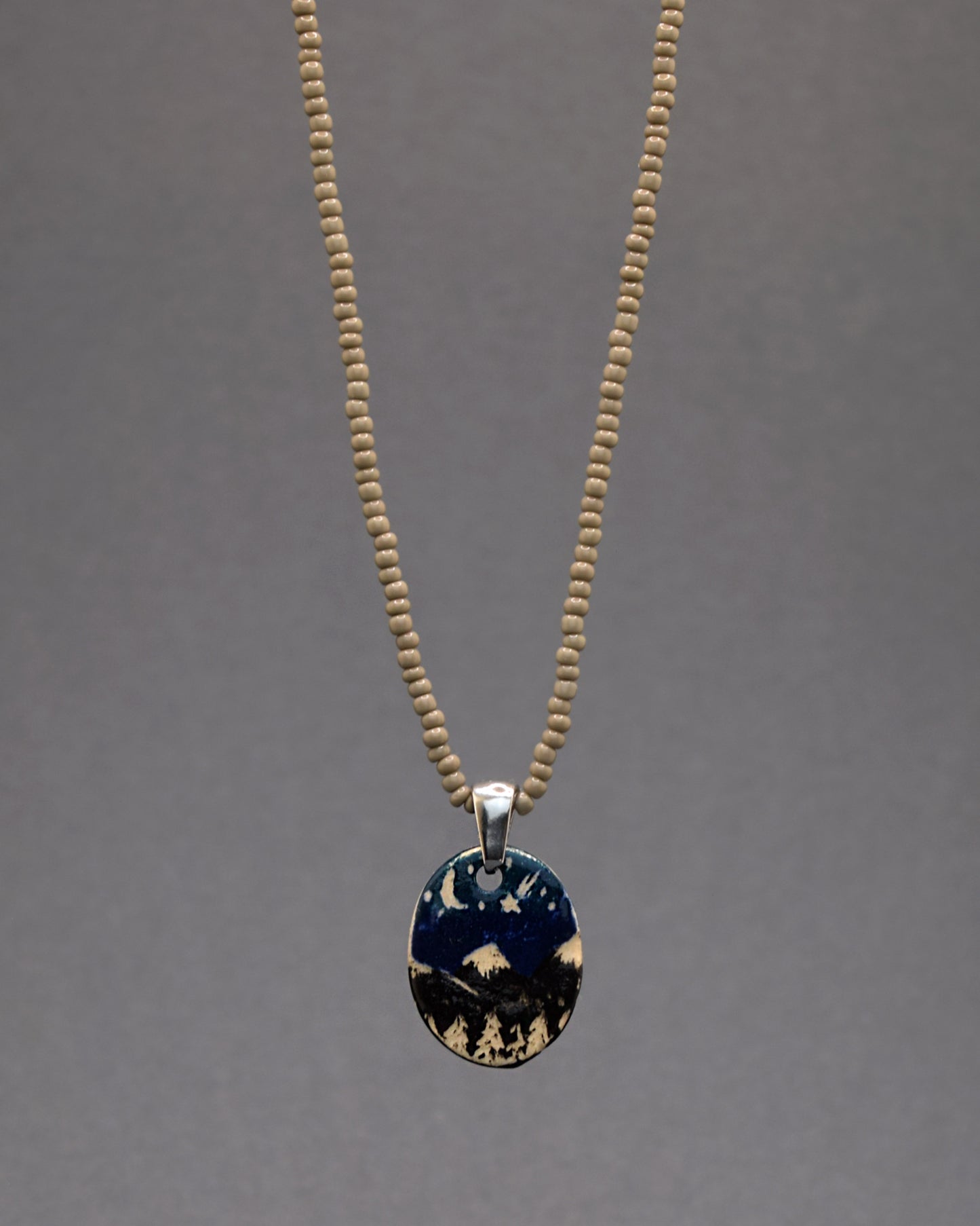 Pendente refinado em cerâmica, meticulosamente pintado à mão, representando uma paisagem de montanhas à noite, suspenso em um colar de missangas de vidro cinzentas. Uma fusão encantadora de arte e elegância, este acessório oferece um toque distintivo de sofisticação e serenidade ao seu visual.