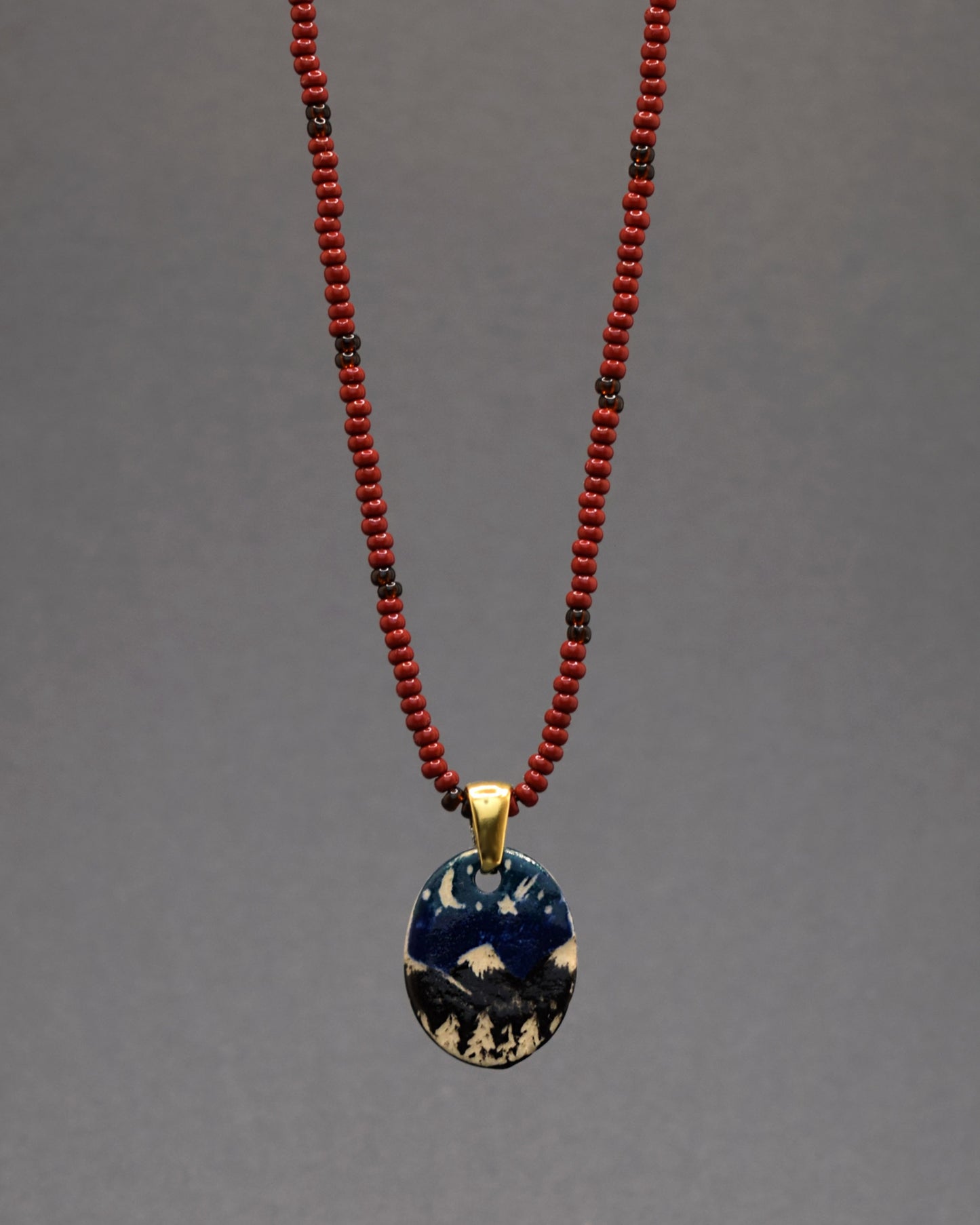 Pendente encantador em cerâmica, minuciosamente pintado à mão, retratando uma paisagem noturna de montanhas. Suspenso em um colar de missangas de vidro bordô, adornado com um padrão gracioso de missangas castanhas, esta peça única combina elegância artesanal e riqueza de cores, adicionando um toque refinado e aconchegante à sua coleção de joias.