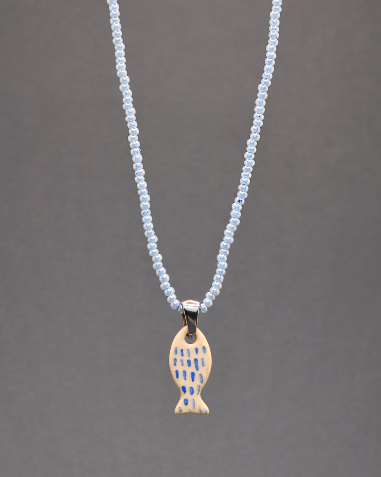 Colar cativante com missangas de vidro azuis claras, realçado por um peixe em cerâmica, meticulosamente pintado à mão com pinceladas em azul que mimetizam as escamas. Uma fusão encantadora de tons suaves e detalhes artísticos, este acessório é uma expressão elegante da beleza natural.