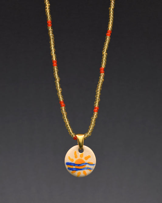 Pendente solar em cerâmica pintada à mão destaca-se com elegância num colar de missangas de vidro douradas com padrão vermelho-alaranjado. Uma fusão exuberante de artesanato refinado e estilo vibrante, esta peça única adiciona um toque sofisticado e caloroso ao seu visual. Celebre a modernidade com um toque de luxo em cada detalhe.