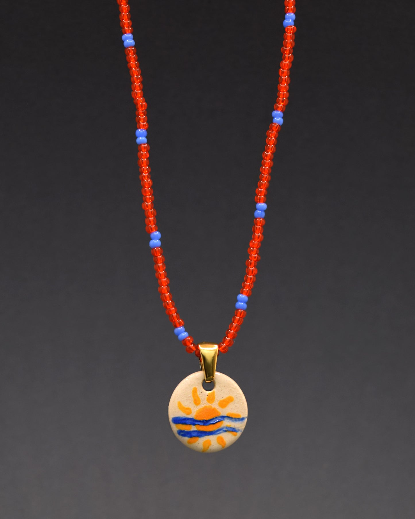 Pendente solar em cerâmica pintada à mão destaca-se com estilo num colar de missangas de vidro vermelho-alaranjado com padrão azul neon. Uma fusão audaciosa de artesanato artístico e cores vibrantes, esta peça única adiciona um toque moderno e ousado ao seu visual. Celebre a contemporaneidade com uma explosão de cores neste elegante acessório.
