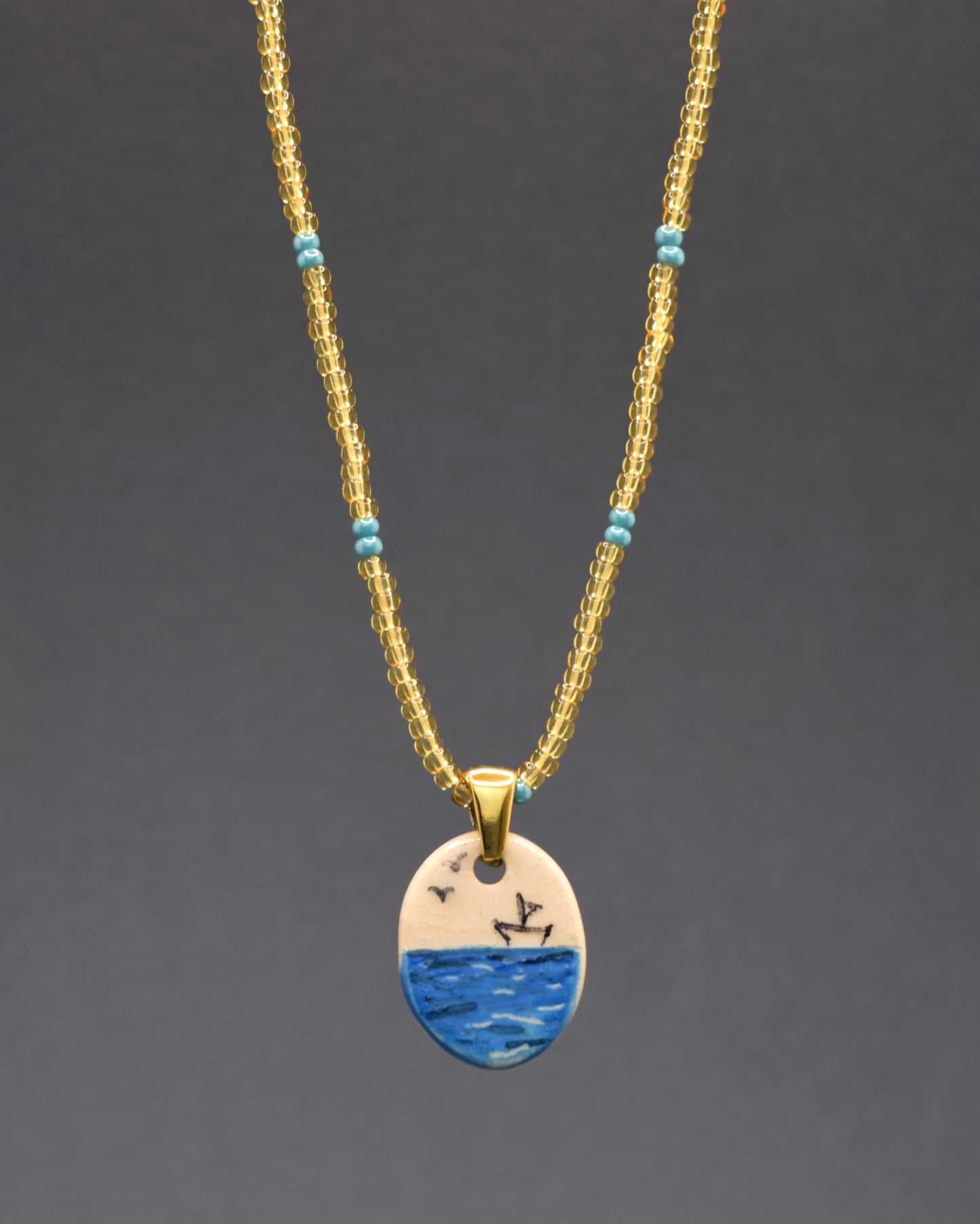 Colar cativante com pendente de cerâmica pintado à mão, retratando um barco de papel no mar. A corrente de missangas de vidro douradas semitransparentes, com padrão azul turquesa, confere uma elegância luminosa à peça. Uma fusão de arte e sofisticação para um visual distintivo.