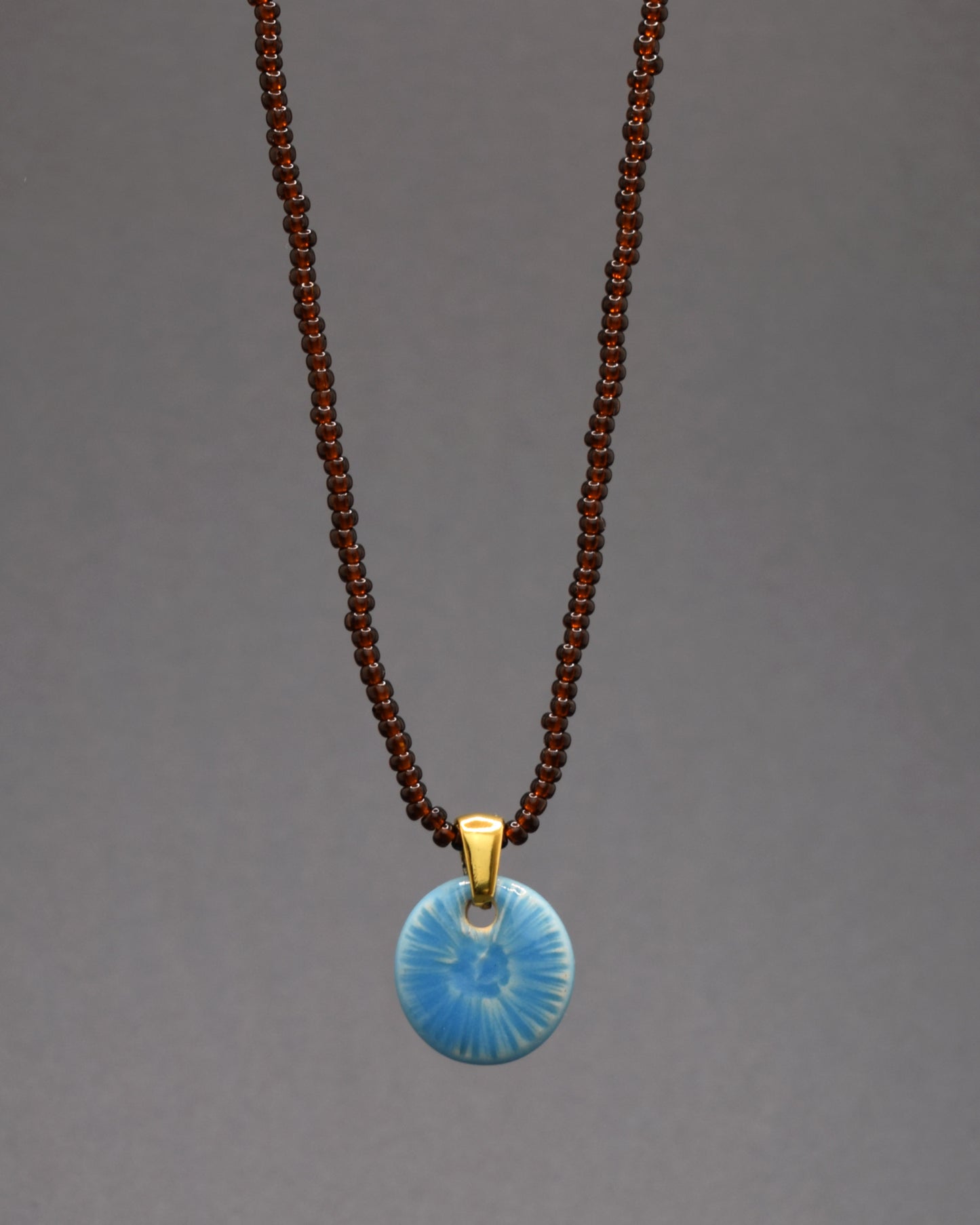 Um colar encantador de missangas de vidro castanhas semitransparentes destaca-se por seu pendente de cerâmica azul celeste, inspirado nas formas delicadas de uma flor do mar. A paleta de tons terrosos transmite serenidade, enquanto o azul celeste adiciona frescor à peça. Uma joia elegante que combina maestria artesanal com a beleza sutil da natureza marinha.