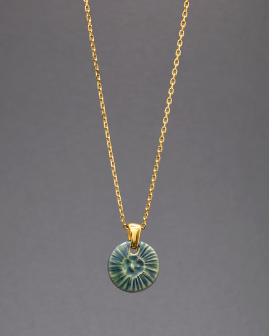 Colar deslumbrante em aço inoxidável dourado, destacando um encantador pendente de cerâmica verde musgo inspirado na beleza de uma flor do mar. A fusão do brilho luxuoso do aço dourado com a serenidade da cerâmica cria uma joia única.