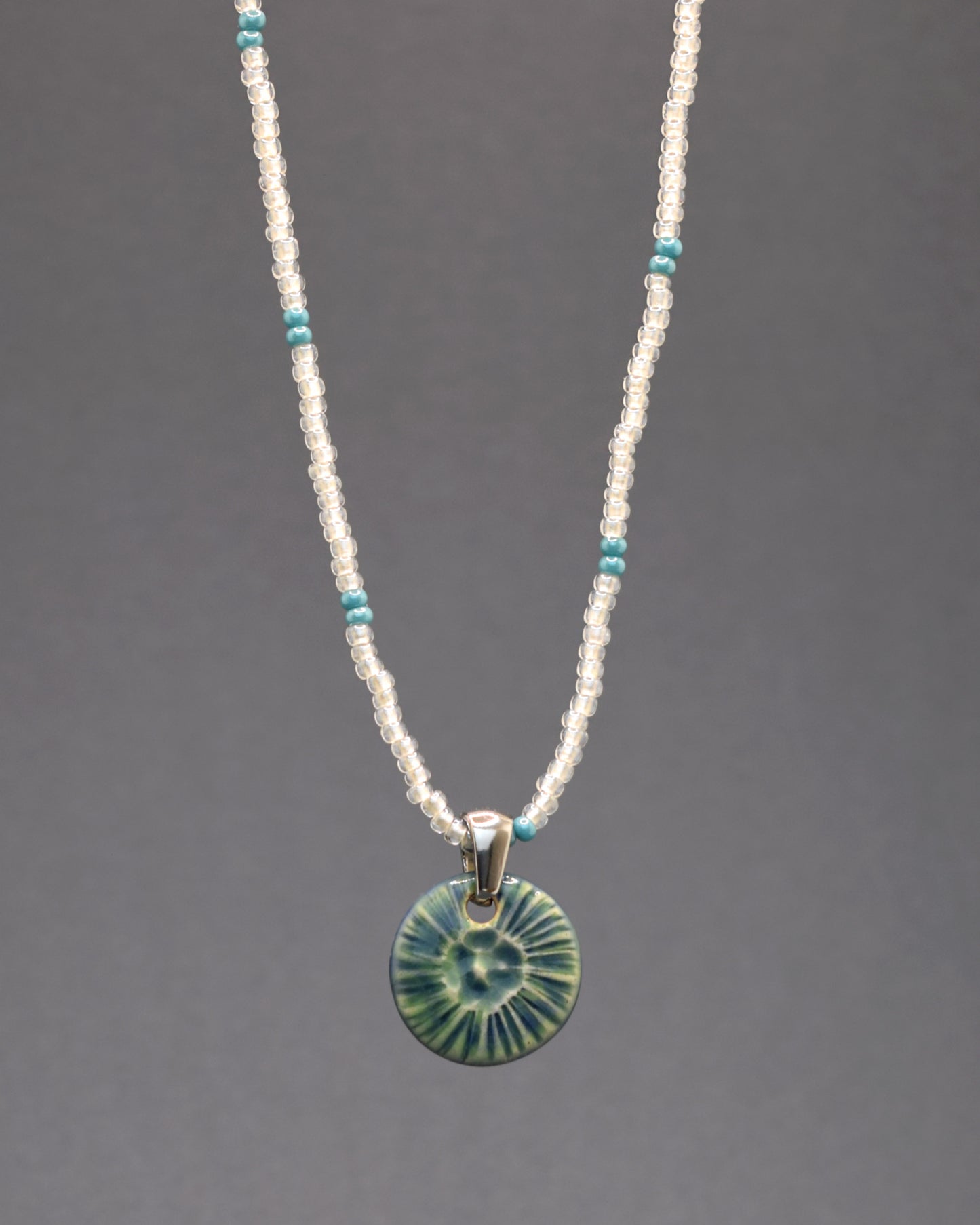 Colar encantador formado por missangas de vidro branco semi-transparente com padrão azul turquesa, acompanhadas por um pendente de cerâmica verde musgo inspirado numa flor do mar. Uma peça distintiva para um toque único ao seu estilo.