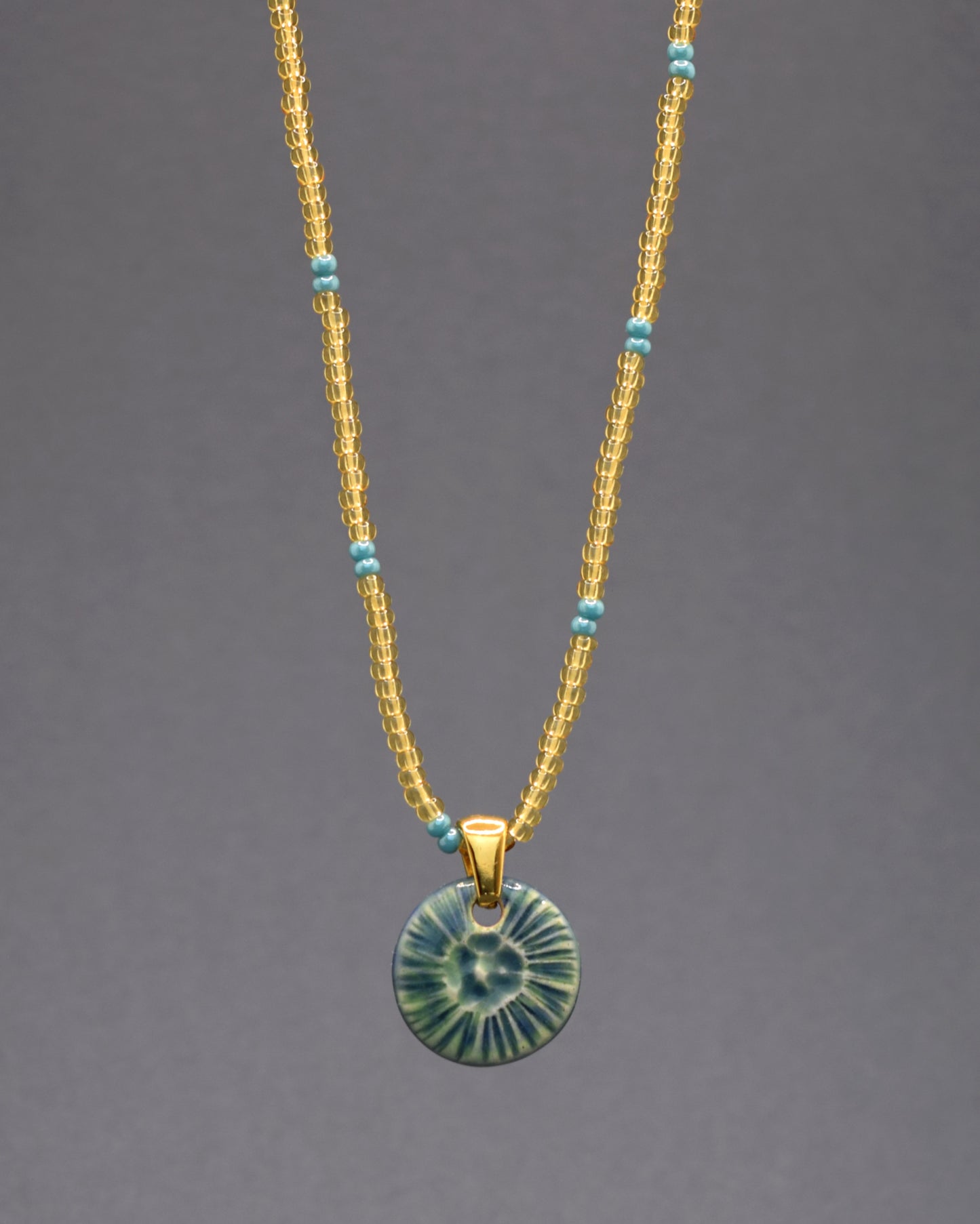 Colar cativante, composto por missangas de vidro dourado semi-transparente, adornadas com um padrão encantador em azul turquesa. O pendente de cerâmica verde musgo, inspirado na beleza de uma flor do mar, completa esta peça distintiva. Uma escolha única para acrescentar um toque exclusivo ao seu estilo.