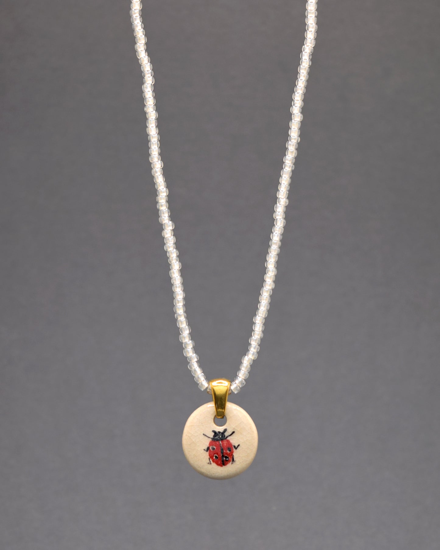 Explore a delicadeza única do nosso colar com pendente de joaninha em cerâmica, meticulosamente pintada à mão, adornando missangas de vidro brancas semitransparentes. Este colar é um convite à pureza e elegância, onde cada detalhe reflete a beleza sutil e a graça intemporal.