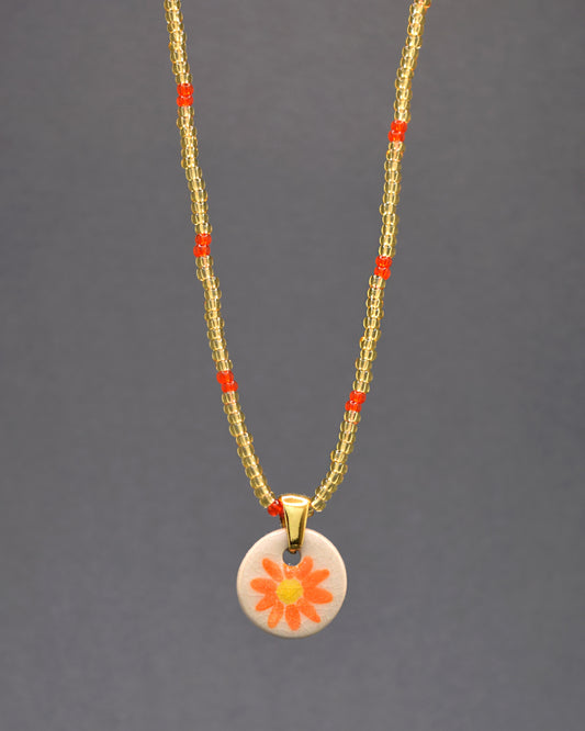 Cative olhares com este colar único! As missangas de vidro douradas semitransparentes com um padrão vermelho-alaranjado, emprestam um brilho sofisticado. O pendente de cerâmica exibe uma margarida laranja, pintada à mão, adicionando um toque artístico e elegante. Um acessório que une o luxo das cores douradas à beleza natural da flora.