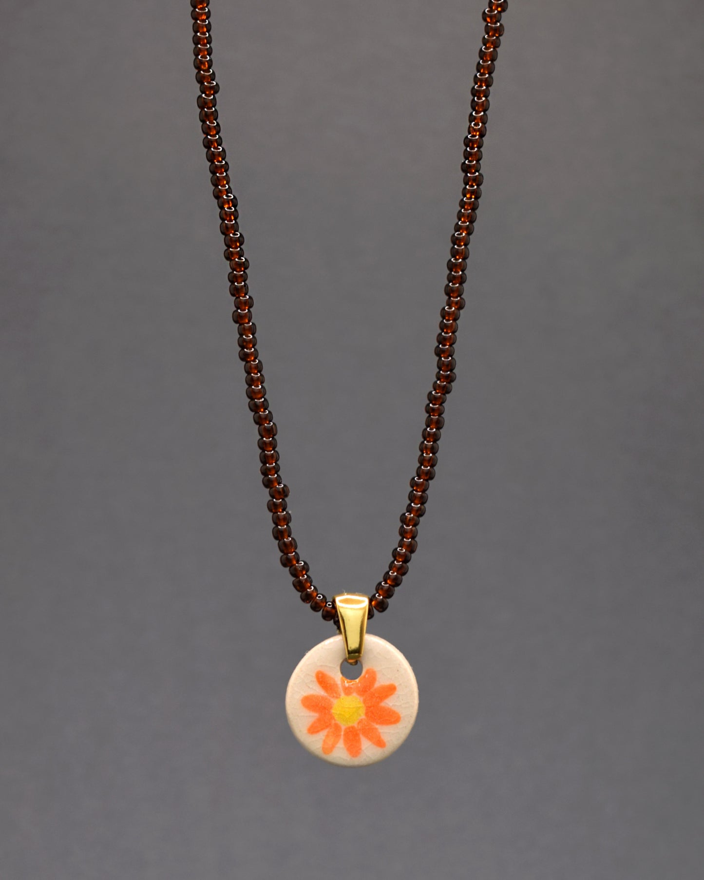 Embrace a beleza natural com este encantador colar. As missangas de vidro semitransparentes, em tons castanhos, criam uma paleta acolhedora. O toque artístico vem do pendente de cerâmica, onde uma margarida laranja é pintada à mão, adicionando um elemento floral delicado e vibrante. Uma peça que captura a essência da natureza com estilo e sofisticação.