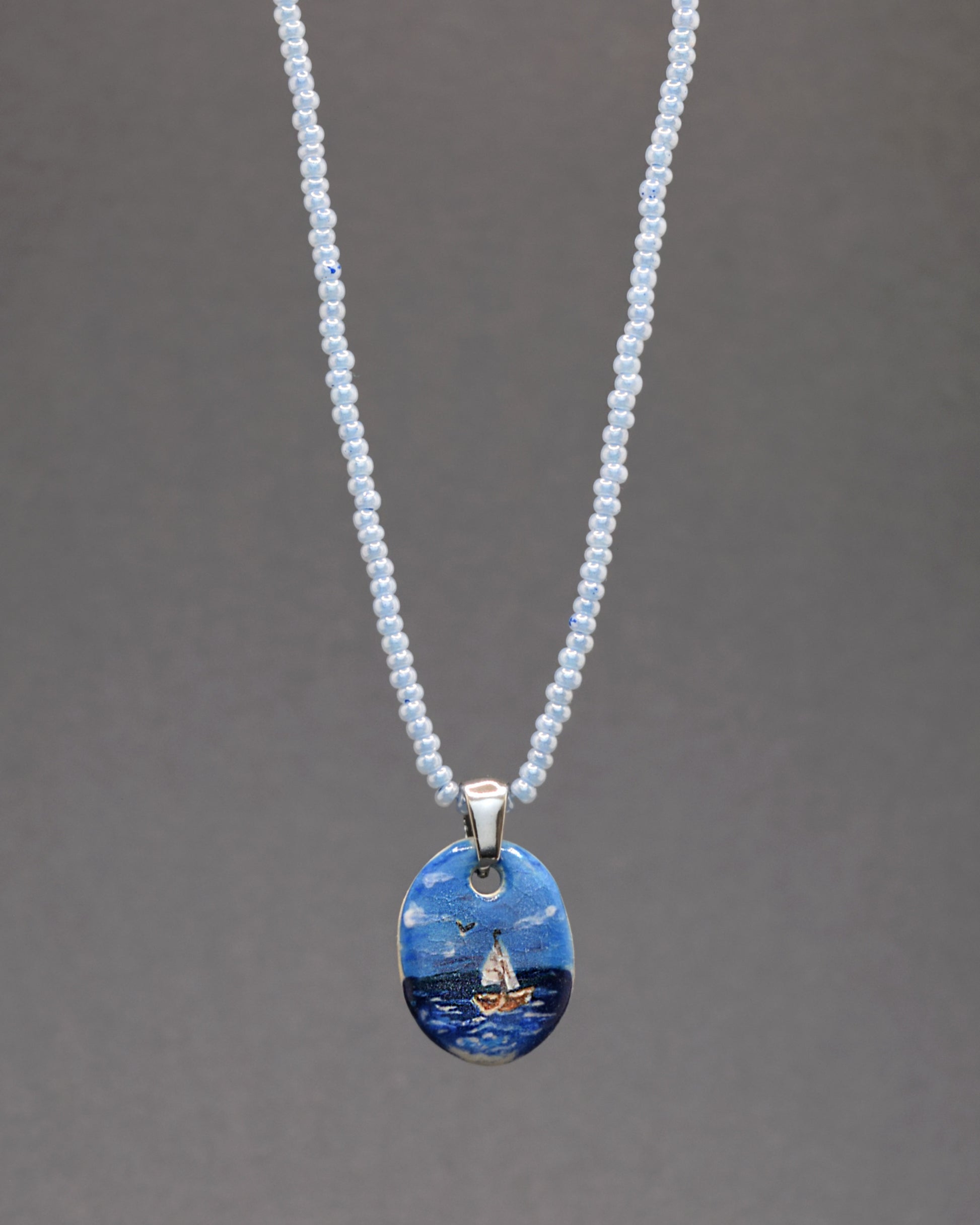Delicadeza em azul! O nosso colar cativante apresenta um encantador pendente de cerâmica, retratando um Barco em Alto Mar, em perfeita harmonia com uma corrente de missangas de vidro azuis claras. Um toque refrescante e elegante para realçar o seu estilo.