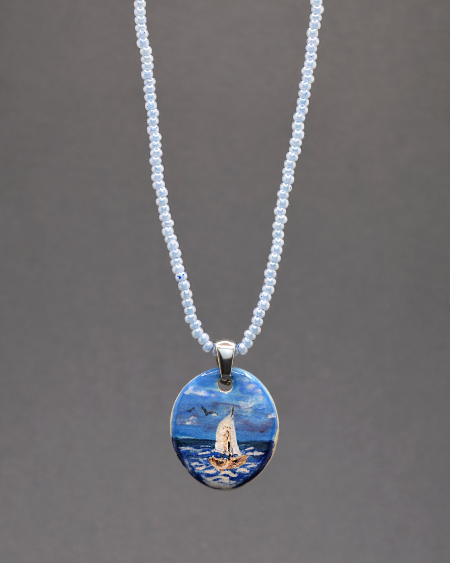 Delicadeza em azul! O nosso colar cativante apresenta um encantador pendente de cerâmica, retratando um Barco em Alto Mar, em perfeita harmonia com uma corrente de missangas de vidro azuis claras. Um toque refrescante e elegante para realçar o seu estilo.