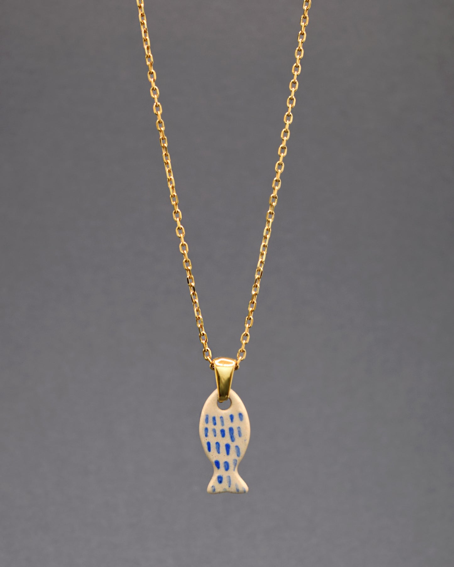 Este colar, com a sua corrente em aço inoxidável dourado, apresenta um charmoso pendente de peixe em cerâmica, pintado à mão com nuances em azul que imitam escamas. Uma fusão perfeita de elegância e arte.