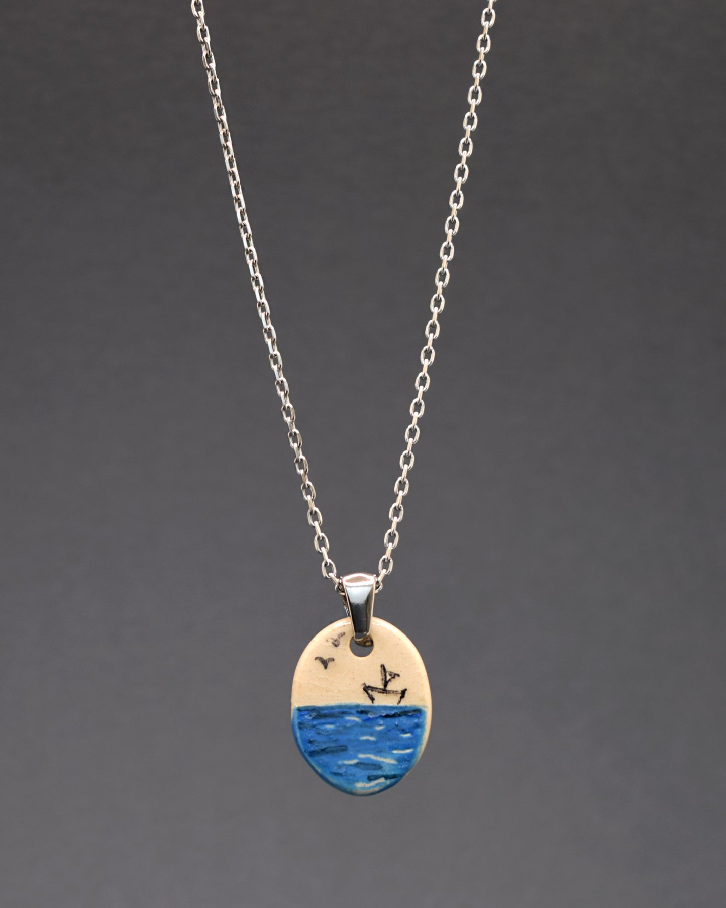 Um colar de aço inoxidável com um toque artístico. Destaca-se o pendente de cerâmica pintado à mão, retratando um barco de papel no mar. Uma fusão de elegância durável e arte delicada em uma única peça.