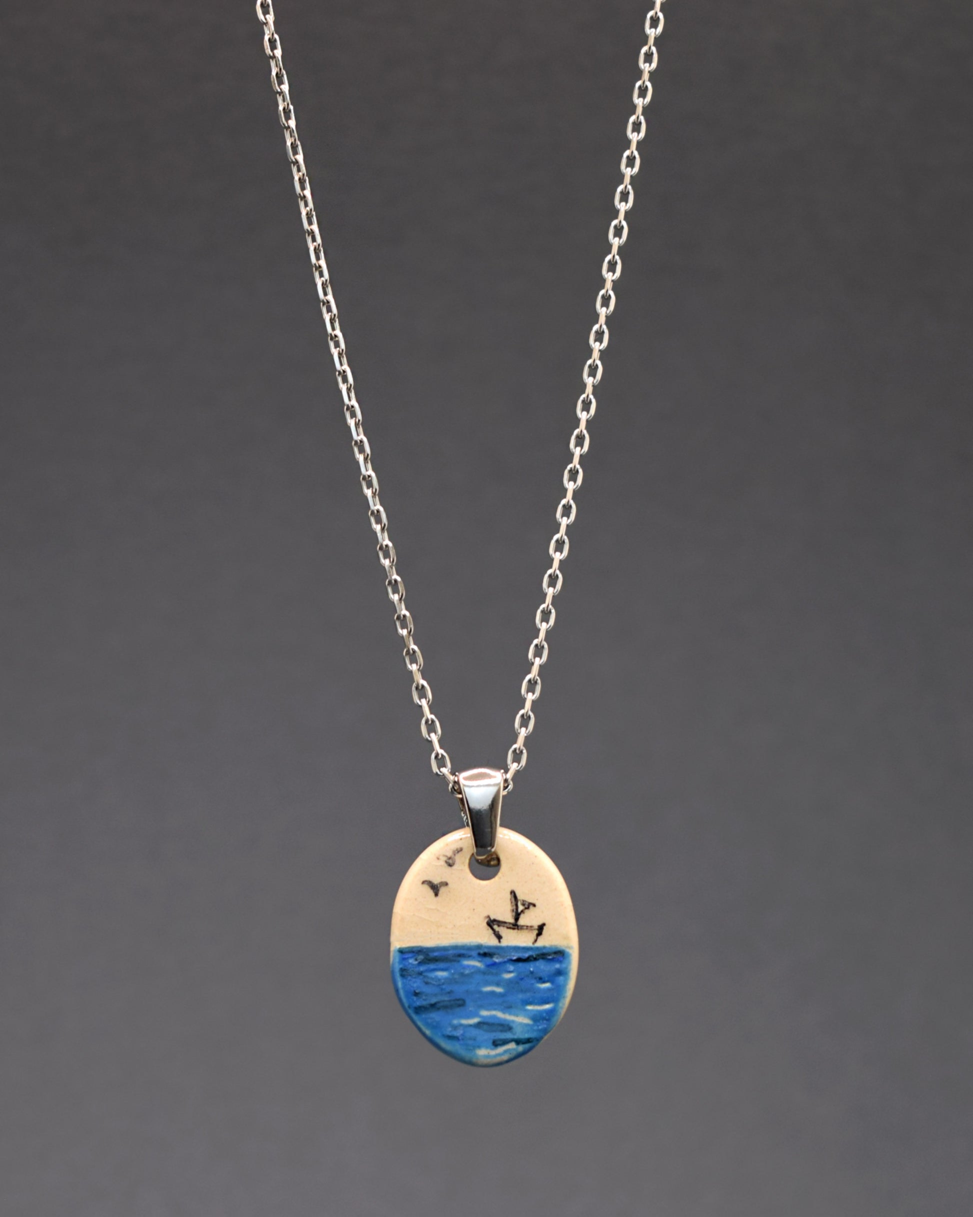 Um colar de aço inoxidável com um toque artístico. Destaca-se o pendente de cerâmica pintado à mão, retratando um barco de papel no mar. Uma fusão de elegância durável e arte delicada em uma única peça.